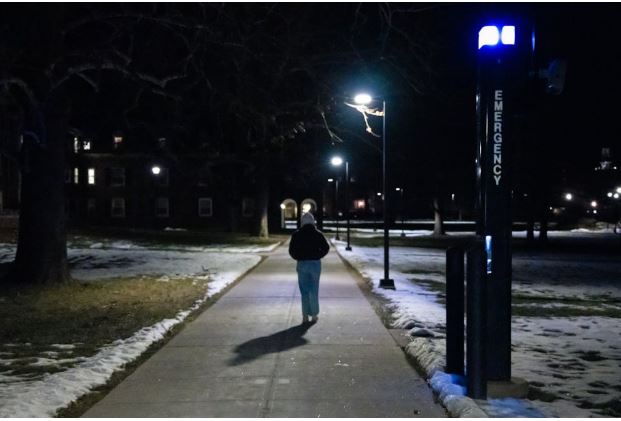 A student walks alone down a lit campus path at night, past a blue-light emergency call station, with snow patches on the ground and dorm buildings in the background.