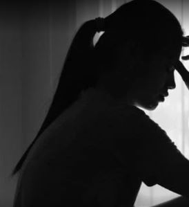 Silhouette of a woman with a ponytail sitting by a window, head resting on her hand in a pensive pose, dramatic black-and-white lighting.