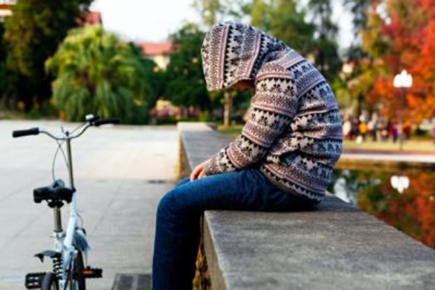 Person in a patterned hooded sweater sitting on a stone ledge with head bowed, bicycle parked nearby beside a park pond in autumn.